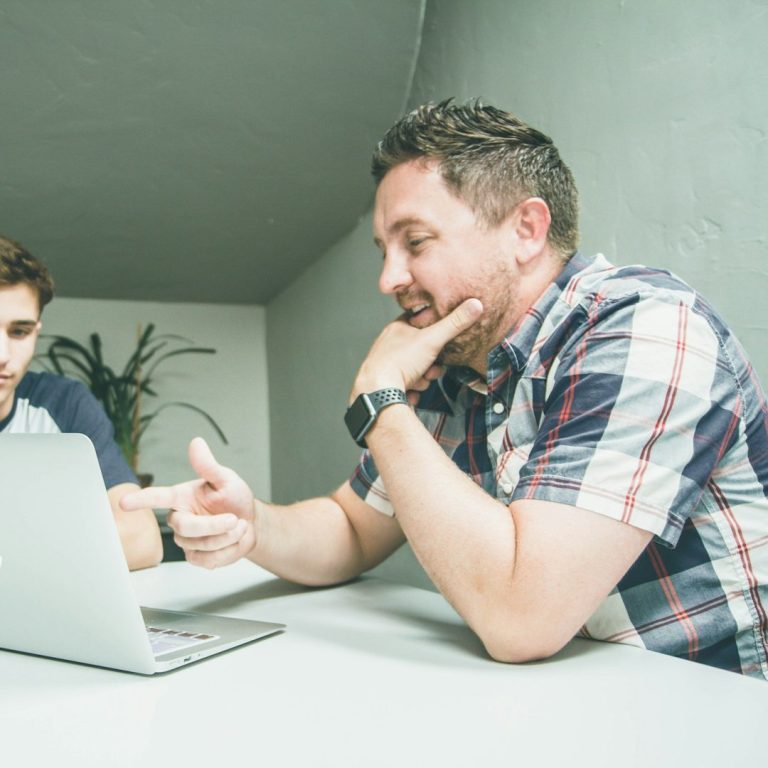 Deux hommes (un jeune et un moins jeune) discutent devant un ordinateur portable dans un cadre intérieur moderne.