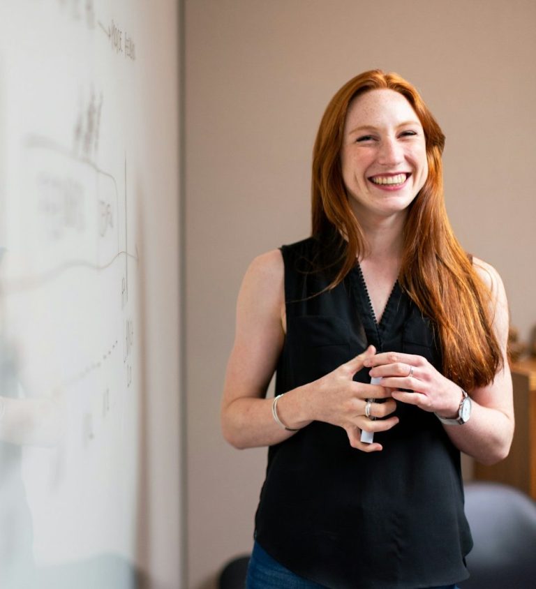 Femme souriante présentant des informations devant un tableau, en conversant avec une autre personne.